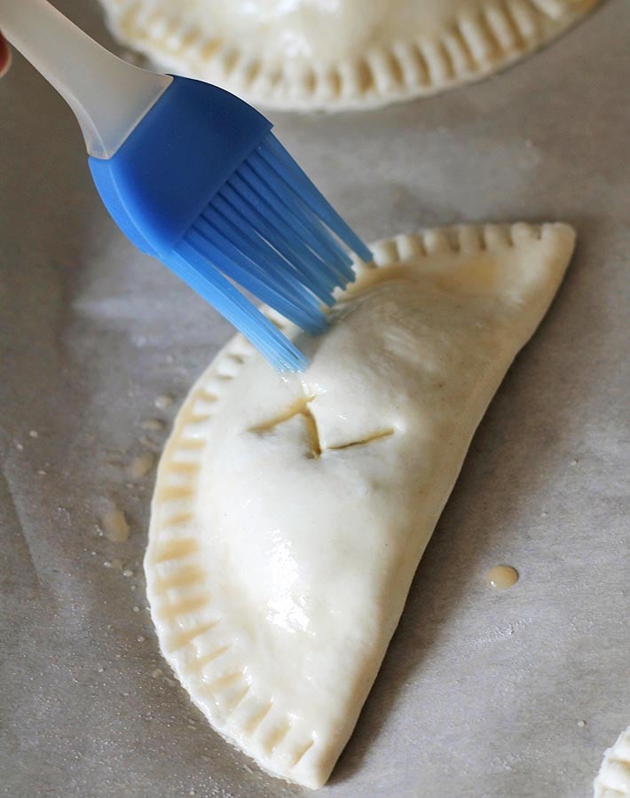 Empanadas de calabaza crudas en una bandeja para hornear forrada con pergamino, una se cepilla con un lavado de huevo vegano justo antes de entrar en el horno.