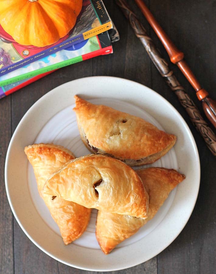 Overhead shot of four vegan pumpkin pasties on a white plate with magic wands sitting off to the right and three Harry Potter books are behind the plate.