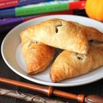 Four pumpkin pasties sitting on a white plate with magic wands in front of the plate and three Harry Potter books sitting in the background.