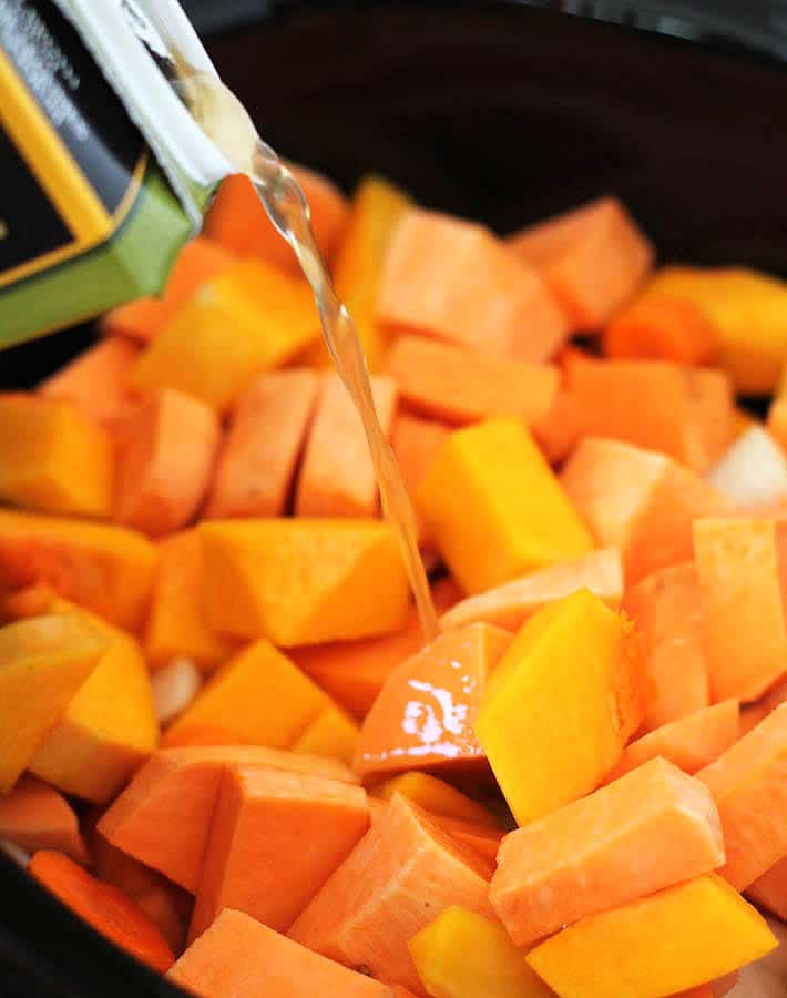 Cubes of sweet potatoes, butternut squash, carrots and onions in a slow cooker with broth being poured over top to make Butternut Squash Sweet Potato Carrot Soup.