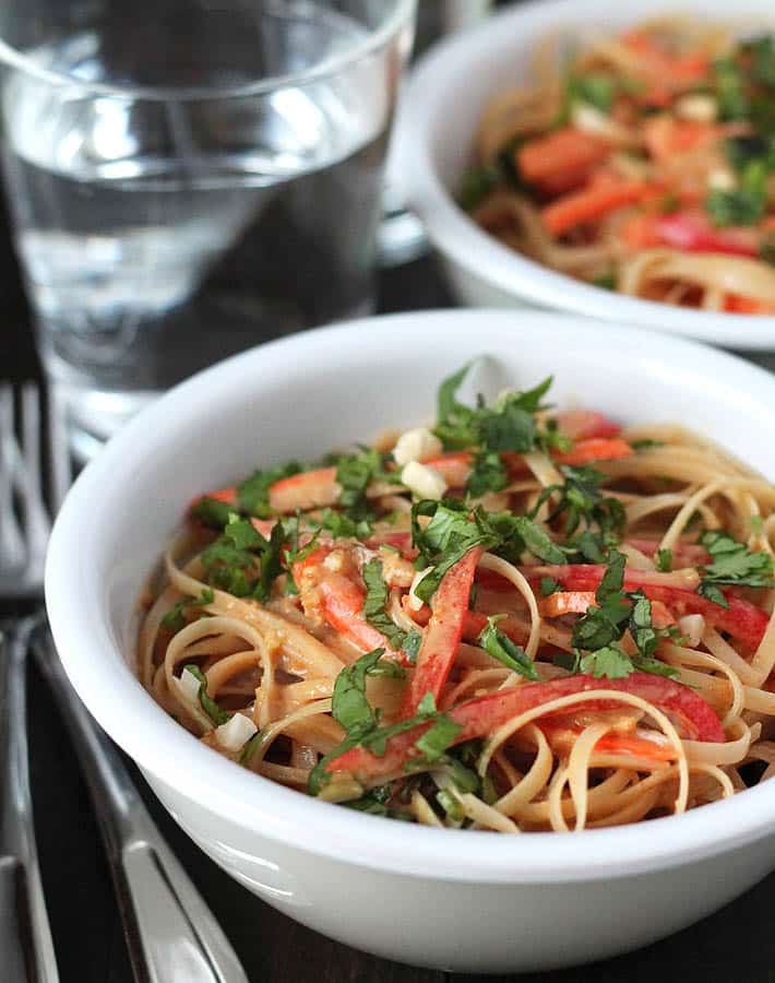 Two bowls of vegan peanut noodles sitting on a dark wooden table with two forks and two glasses of water to the left.