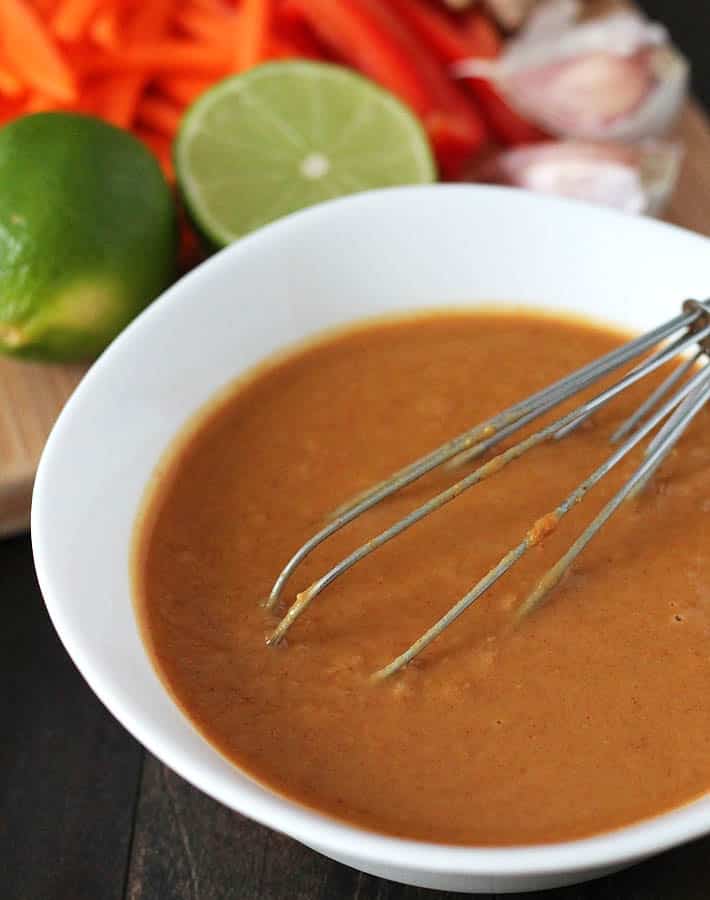 A small white bowl filled with freshly made peanut sauce for vegan peanut noodles and a small wire whisk is sitting in the bowl and the ingredients used to make the sauce are sitting behind the bowl on a bamboo cutting board.