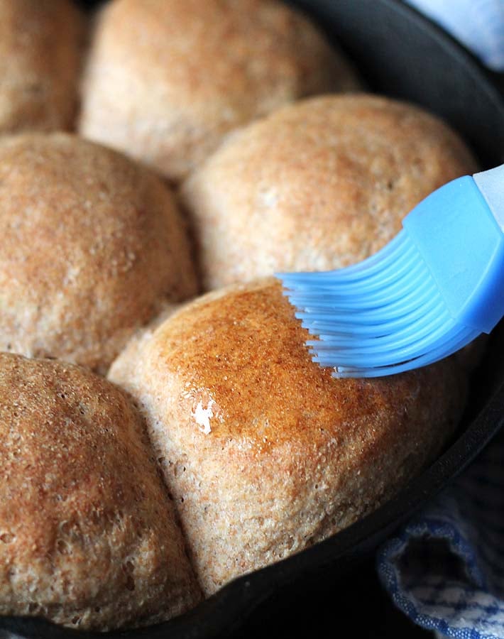 A cast iron pan of spelt rolls just out of the oven being brushed with butter with a blue silicone brush.