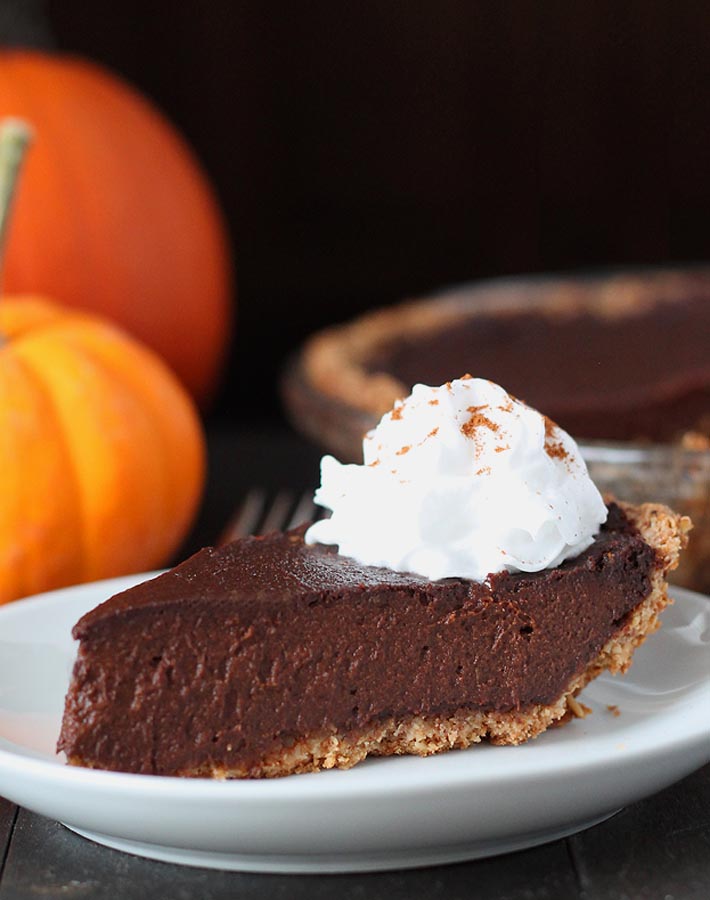 An close up shot of a slice of vegan chocolate pumpkin pie on a plate with the whole pie, minus one slice, behind to the right and fresh pumpkins behind to the left.