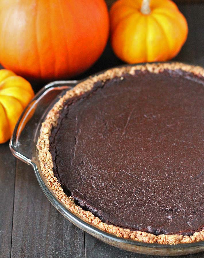 A whole vegan chocolate pumpkin pie just out of the oven, sitting on a black wooden table with pumpkins behind the pie.