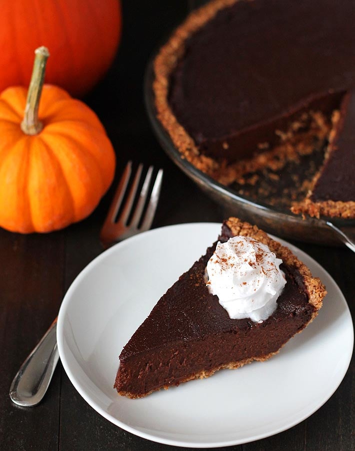 A slice of vegan chocolate pumpkin pie sitting on a small white plate with coconut whipped cream garnish, with a fork to the left and the rest of the pie to the right.