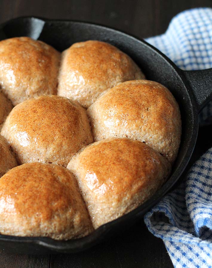 Fresh spelt rolls in a cast iron pan that have been brushed with butter and a blue dish cloth sitting to the right.