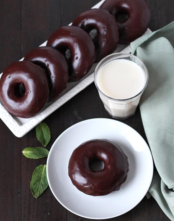 An overhead shots of Vegan Gluten Free Chocolate Mint Doughnuts on a white rectangle plate and one doughnut sitting on a small white plate with a glass of almond milk to the right of the small white plate and three fresh mint leaves sitting to the left of the white plate.