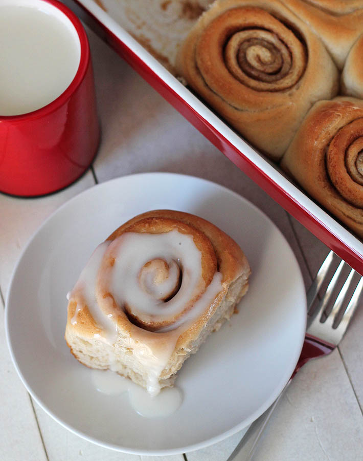 Overhead shot of Easy Vegan Cinnamon Rolls in a red baking dish with a red mug of almond milk to the left side of the dish and one roll sitting on a small white plate in front of the mug.