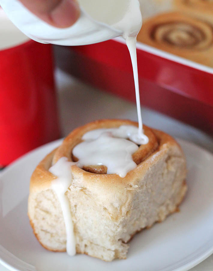 Easy Vegan Cinnamon Rolls in a baking dish with icing being drizzled over one sitting on a small white plate in front of the baking dish.