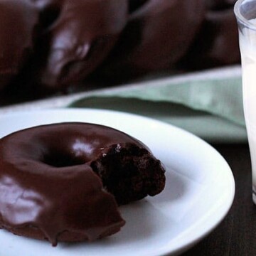 A row of Vegan Gluten Free Chocolate Mint Doughnuts on long rectangle plate in the background and one doughnut with a bite taken out of it sitting on a small white plate with a glass of almond milk to the right.