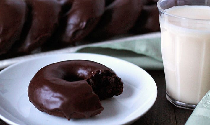 A row of Vegan Gluten Free Chocolate Mint Doughnuts on long rectangle plate in the background and one doughnut with a bite taken out of it sitting on a small white plate with a glass of almond milk to the right.