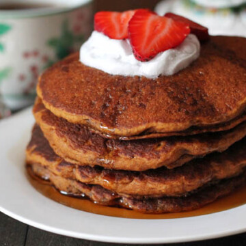 Four Gluten Free Vegan Gingerbread Pancakes stacked on a white plate and topped with coconut whipped cream and fresh slices of strawberries.