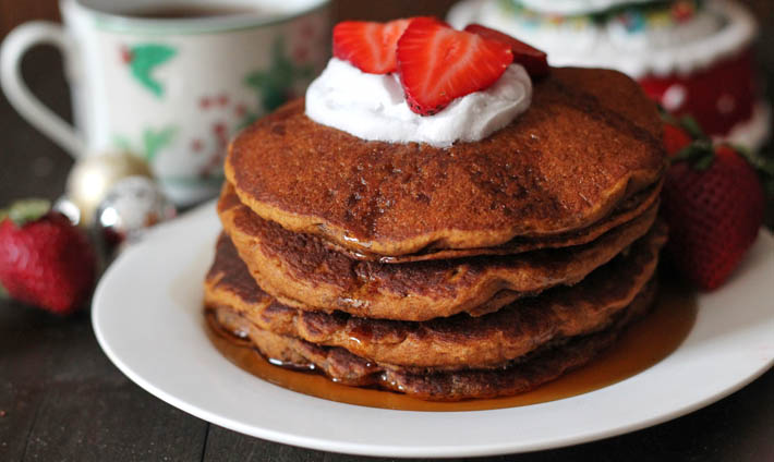 Four Gluten Free Vegan Gingerbread Pancakes stacked on a white plate and topped with coconut whipped cream and fresh slices of strawberries.