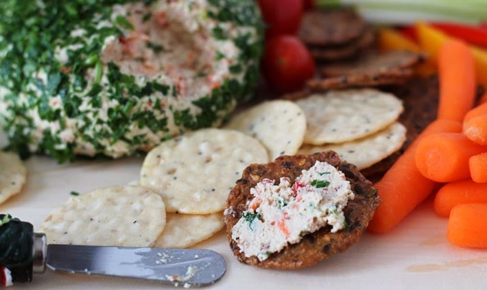 A cracker with some Vegan Vegetable Almond Cheese Ball spread on it, cracker is sitting on a board with the cheese ball behind it and cut up vegetables surround the cheese ball.