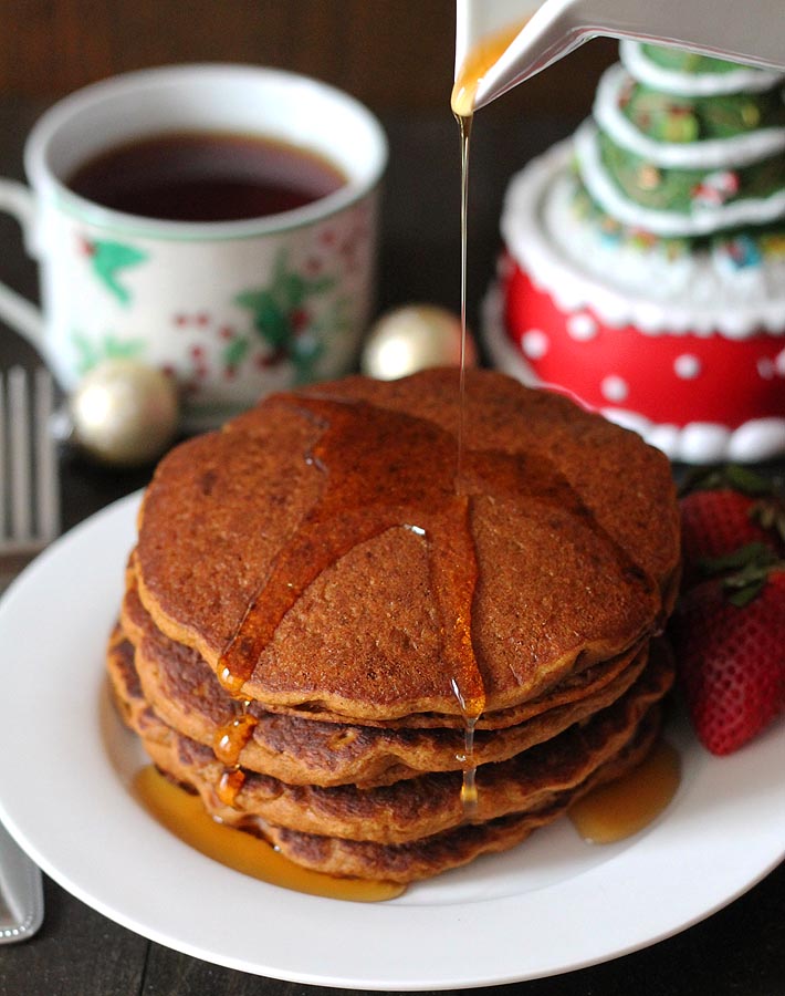 A stack of Gluten Free Vegan Gingerbread Pancakes on a white plate with maple syrup being poured over top, a cup of tea sits in the background in a small mug that is decorated with holiday holly's.