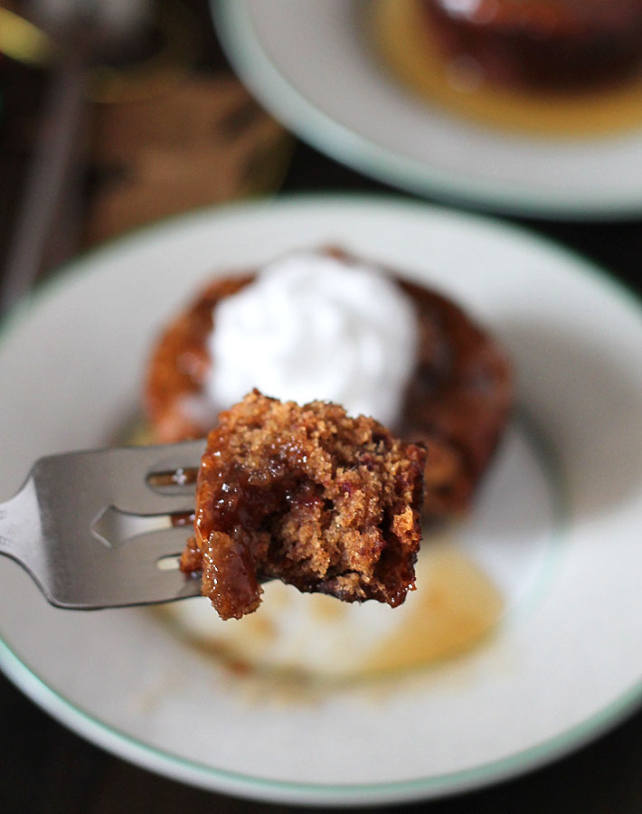 A close up shot of a piece of vegan sticky toffee pudding on a fork, the rest of the pudding is sitting on a plate in the background.