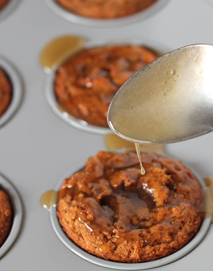 Vegan sticky toffee puddings in a muffin tin, maple caramel sauce is being drizzled onto one of them.