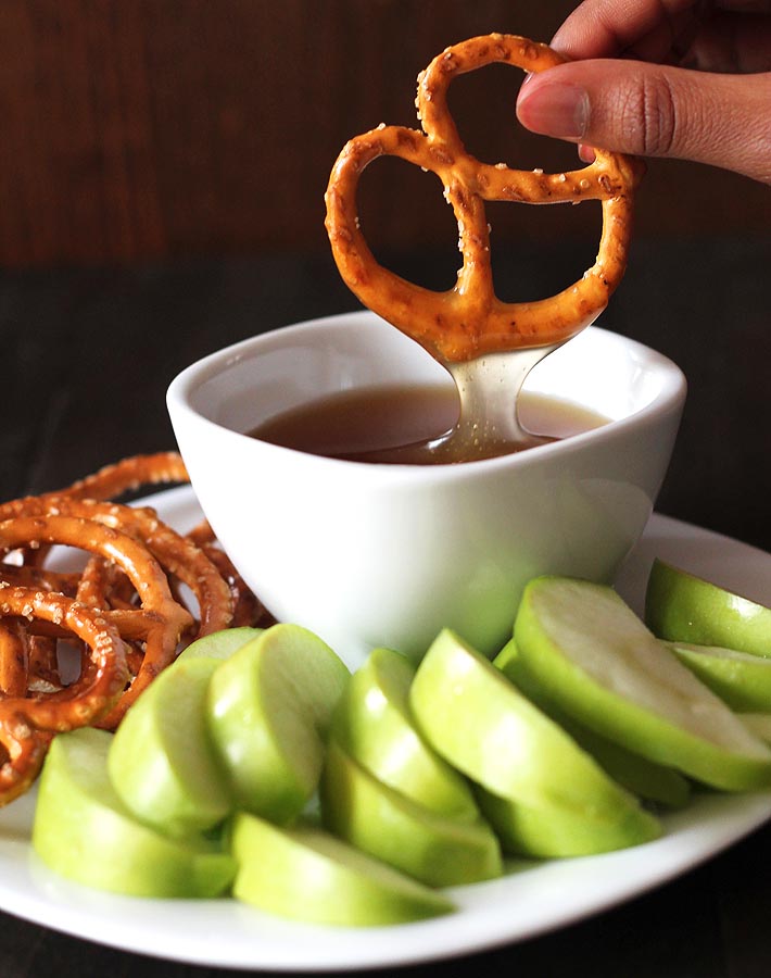 A pretzel just dipped in a bowl of vegan maple caramel sauce with sliced green apples and more pretzels sitting on a plate.