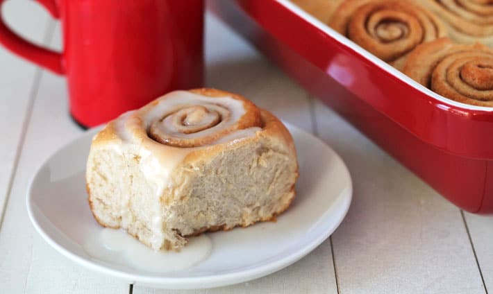 Easy Vegan Cinnamon Rolls sitting in a baking dish with one roll in front of the dish on a small white plate and a red mug filled with almond milk sits behind the plate.