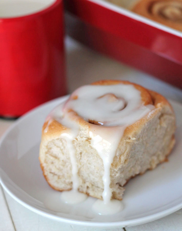 Easy Vegan Cinnamon Rolls in a red baking dish on a white table and one cinnamon roll with icing sits on a small white plate in front of the baking dish.