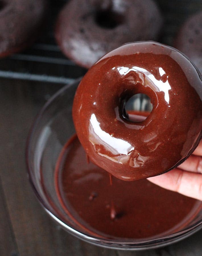 A batch of Vegan Gluten Free Chocolate Mint Doughnuts sitting on a cooling rack in the background and a freshly dipped doughnut being held with glaze dripping off of it.