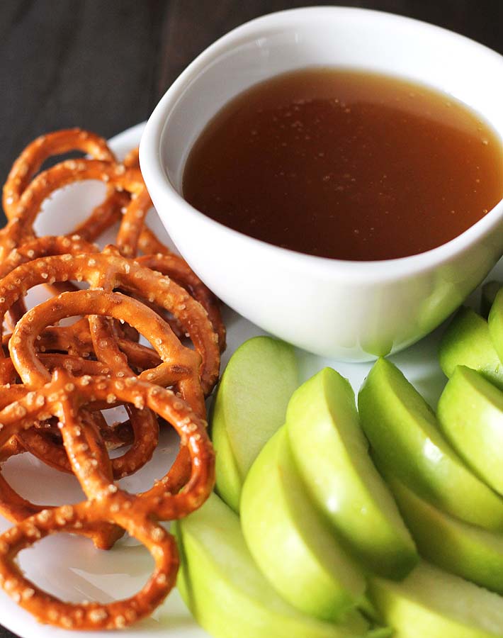 An overhead shot of a white bowl filled with dairy free maple caramel on a white plate with sliced green apples and pretzels.