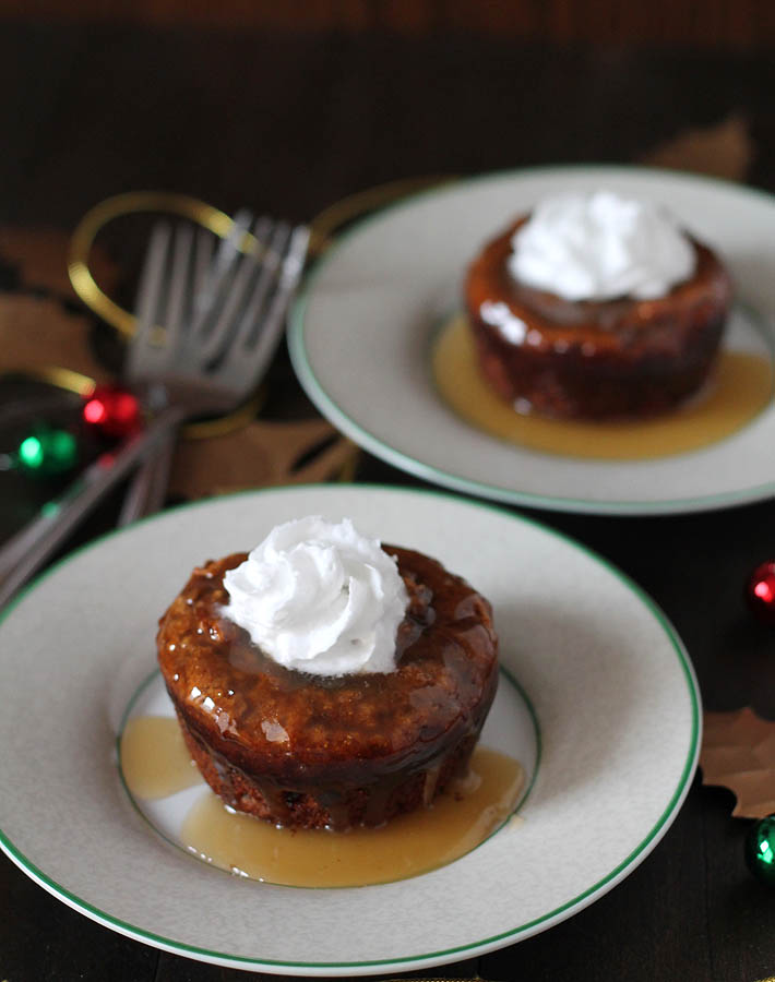 Two plates, each with a vegan sticky toffee pudding sitting on it, two forks are on the left side of the plates and the table they are sitting on is decorated for the holidays.