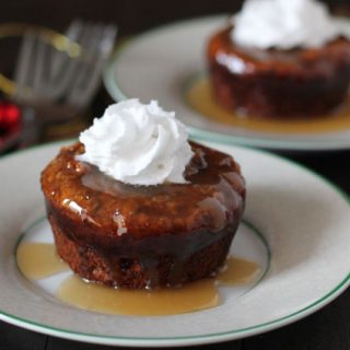 A mini vegan sticky toffee pudding sitting on a small plate, pudding has a small dollop of coconut whipped cream on top of it, two forks sit off to the left of the plate and another plate is in the background.