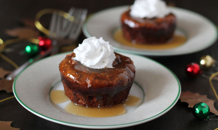 A mini vegan sticky toffee pudding sitting on a small plate, pudding has a small dollop of coconut whipped cream on top of it, two forks sit off to the left of the plate and another plate is in the background.