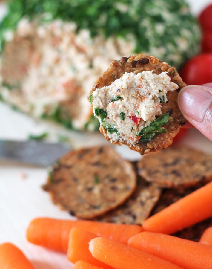 A hand holding a cracker with Vegan Vegetable Almond Cheese Ball spread onto it to show the texture of the veggie filled spread, the rest of the vegan cheese balls sits in the background on a wooden board.