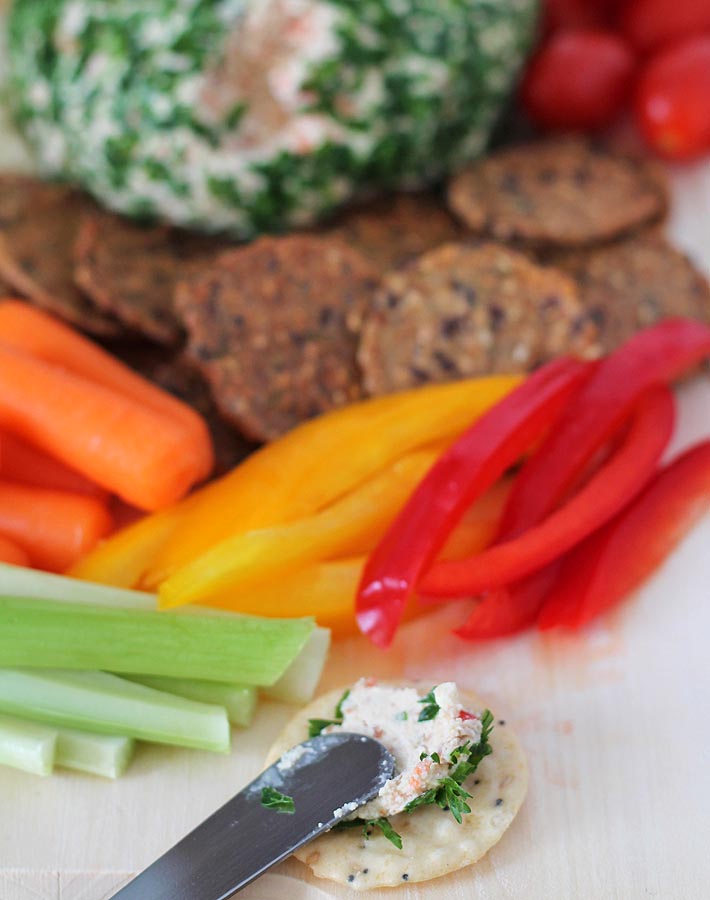 A round rice cracker with a little bit of Vegan Vegetable Almond Cheese Ball spread onto it, the cheese ball sits in the back of the cracker off to the left and veggies sit off to the right behind the cracker.