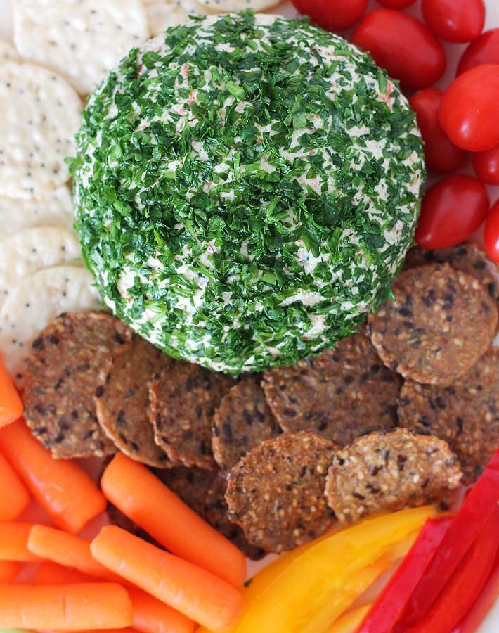 A Vegan Vegetable Almond Cheese Ball rolled in finely chopped parsley sitting on a wooden board surrounded by two types of crackers and baby carrots, cherry tomatoes, and sliced red and yellow peppers.