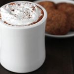 Creamy Vegan Hot Chocolate topped with coconut whipped cream in a white mug with a plate of cookies on a plate behind it.