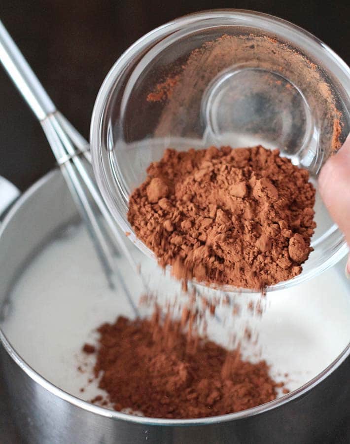 Cocoa powder being poured into a pot with non-dairy milk, a whisk sits in the pot to mix and make Creamy Vegan Hot Chocolate.