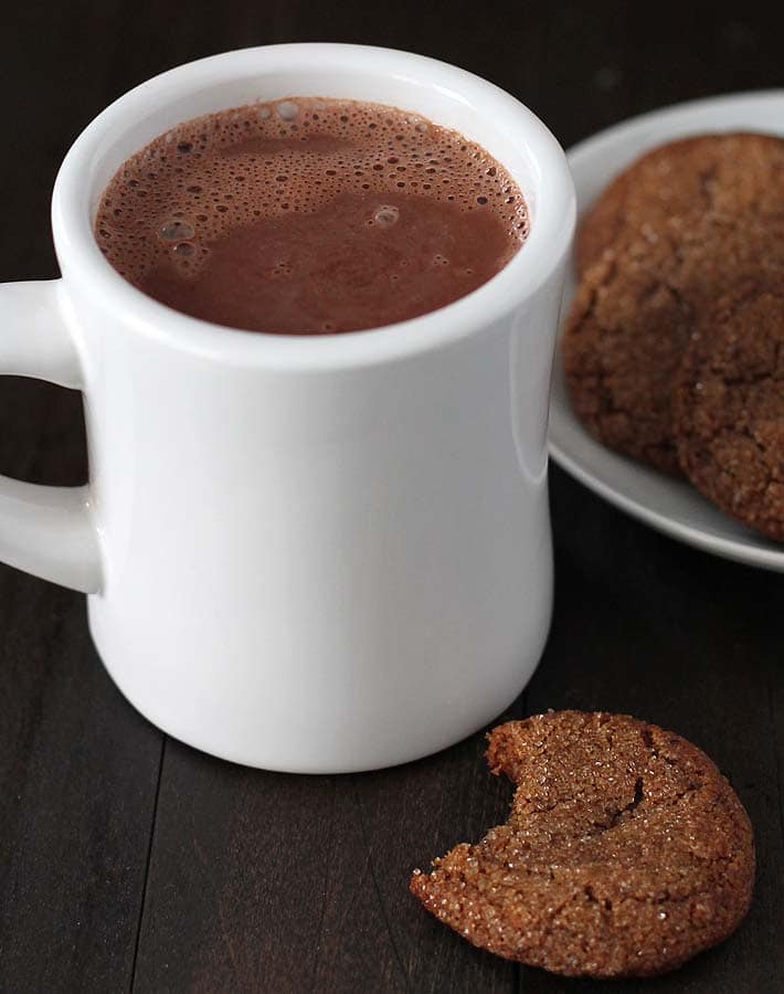 Creamy Vegan Hot Chocolate in a white mug on a dark wood table with a plate of cookies in behind it and a cookie with a bit taken out of it in front of the mug on the table.