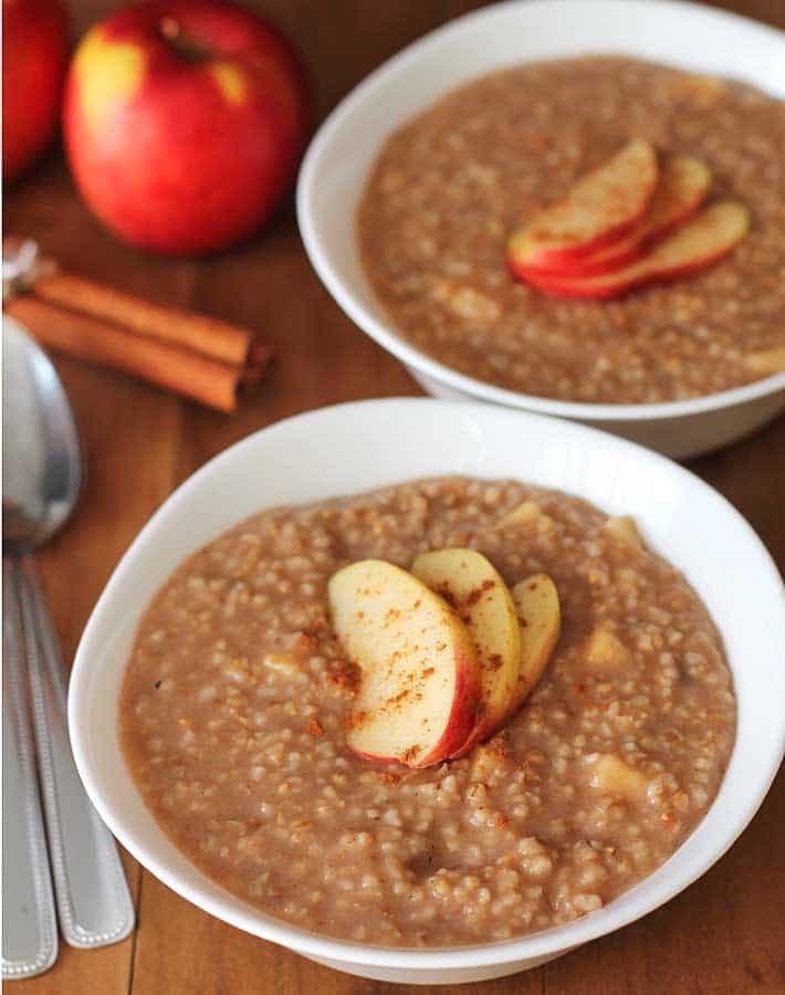 Apple Spice Instant Pot Steel Cut Oats in two white bowls with 3 fresh apple slices on top of each bowl of oat, bowls are sitting on a brown wood table.