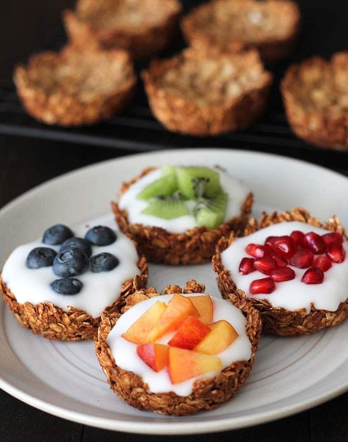 Four granola cups sitting on a plate, each are filled with vegan coconut yogurt, one is topped with fresh blueberries, another with fresh kiwi pieces, another with pomegranate arils, and the last with fresh nectarine pieces.