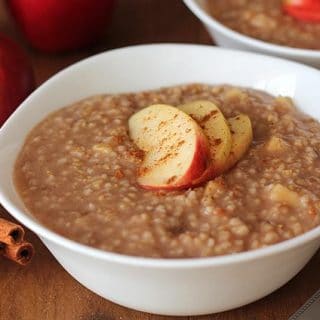 Apple Spice Instant Pot Steel Cut Oats in a white bowl with 3 fresh apple slices on top, three red apples sit to the left of the bowl and cinnamon sticks sit on front of the apples.