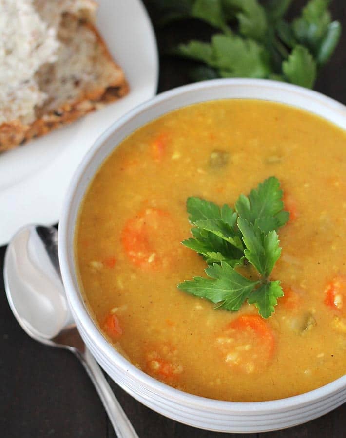 Overhead shot of a bowl of Curried Instant Pot Split Pea Soup in a white bowl, soup is garnished with a small bunch of fresh parsley; a spoon sits to the left of the bowl.