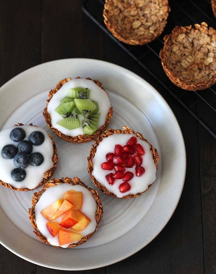 Overhead shot of fruit and yogurt filled granola cups on a white plate, each granola cup is filled topped with a different fresh fruit: pomegranate arils, kiwi pieces, blueberries, and nectarines.