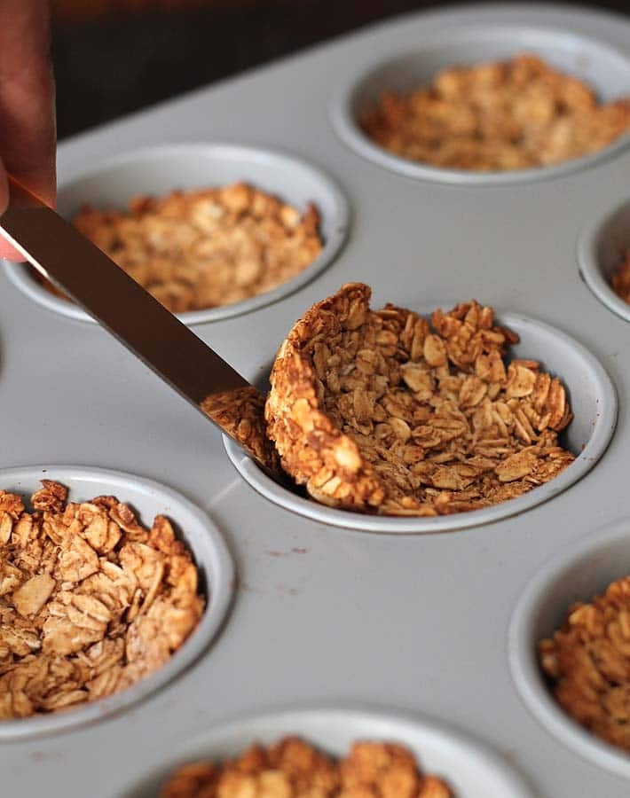 Freshly baked granola cups in a muffin pan, one granola cup is being removed with an icing spatula.