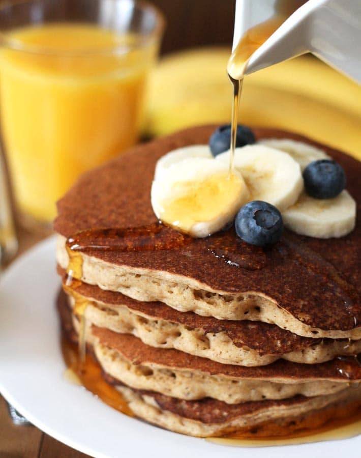 Maple syrup being poured from a white ceramic cup onto four Vegan Banana Pancakes that are sitting on a white plate.