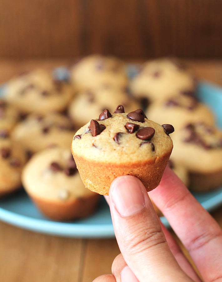 A blue plate of Mini Chocolate Chip Muffins sits in the background on a brown wood table, in the forefront of the image, a hand is holding up a muffin.