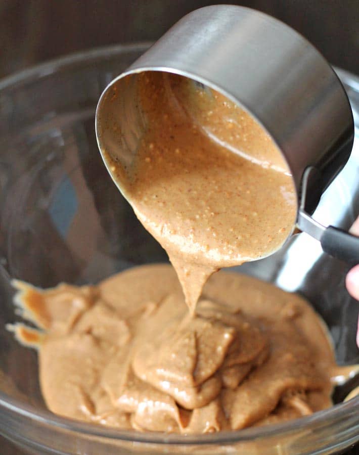 Natural peanut butter bring poured from a measuring cup into a glass mixing bowl to make Peanut Butter Oatmeal Chocolate Chip Cookies.