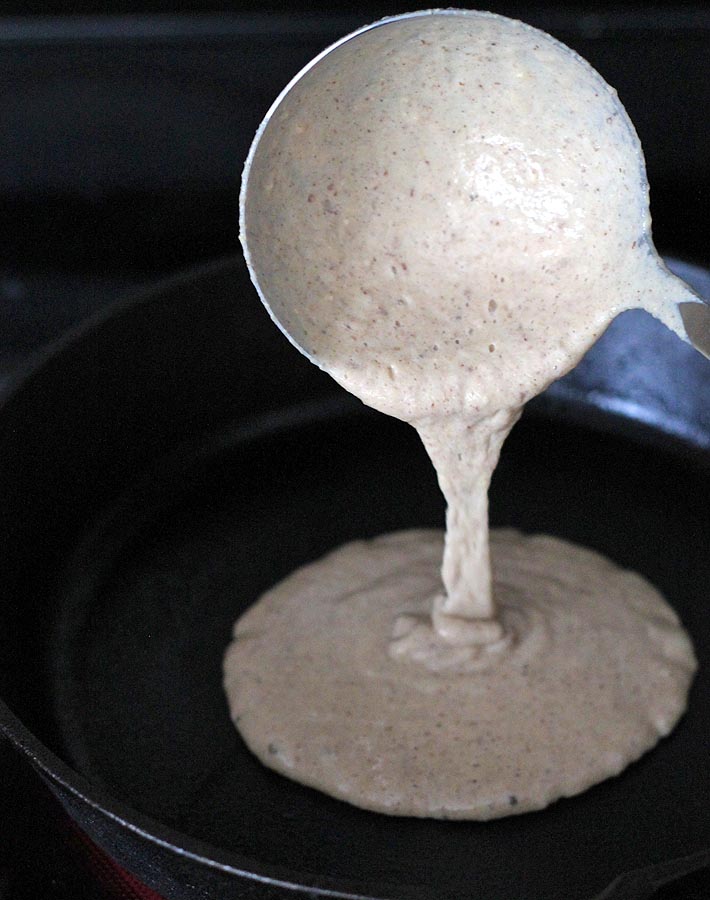 Batter for Vegan Banana Pancakes being poured by a stainless steel ladle into a cast iron pan.
