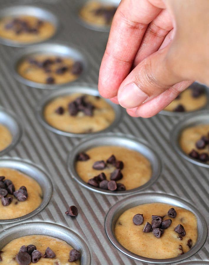 Batter for Mini Chocolate Chip Muffins portioned into a mini muffin pan, a hand is sprinkling mini chocolate chips onto each muffin.