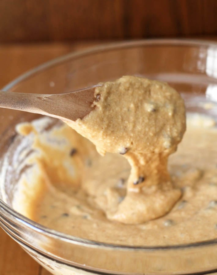 The batter for mini chocolate chip muffins dropping from a wooden spoon into a glass bowl.