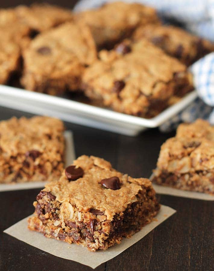 Three Chocolate Chip Peanut Butter Oatmeal Bars on a dark wood table, a plate of more bars sits in the background.