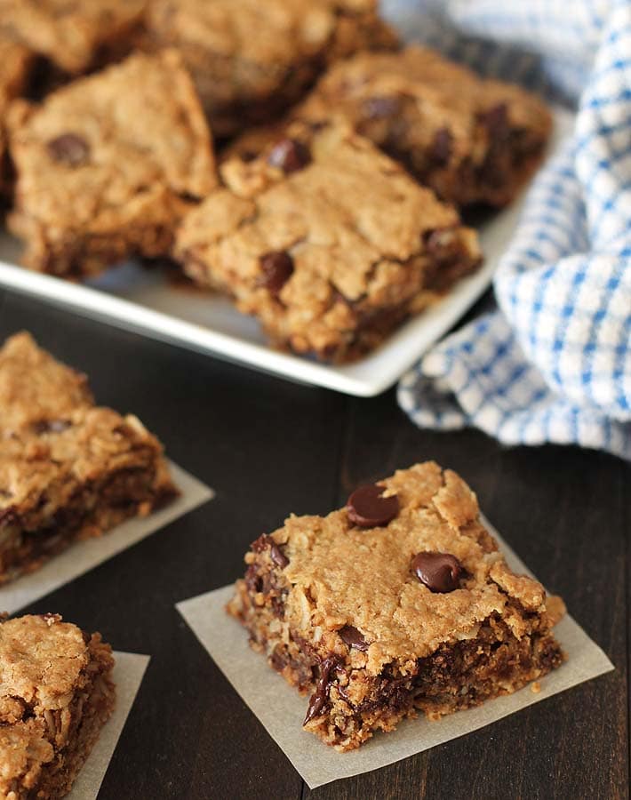 Chocolate Chip Peanut Butter Oatmeal Bars on parchment paper squares sitting on a dark wood table, a white plate of more bars sits in the background.